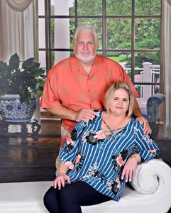 Fernie and Sue Soignier posing next to a white couch in front of a large window looking out into a landscaped front lawn.